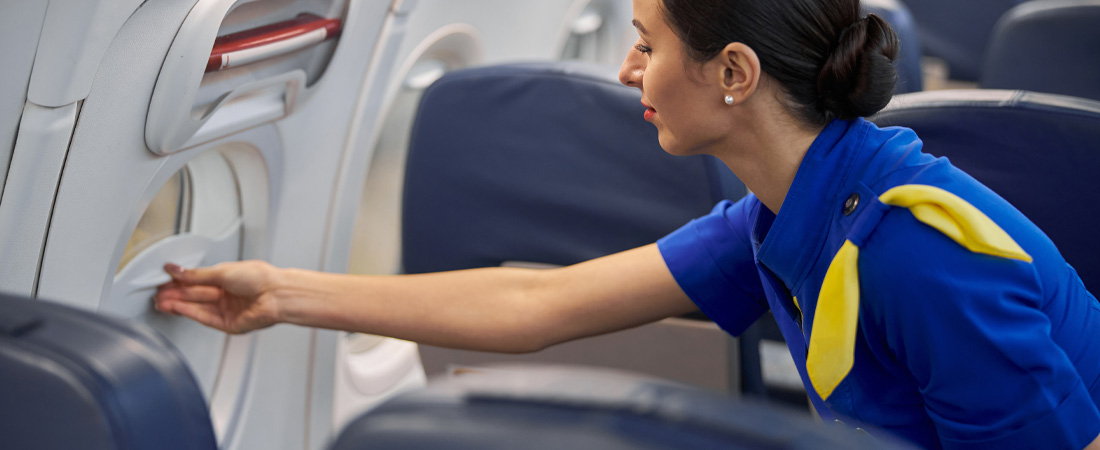 Primer plano de una asistente de vuelo con uniforme, cerrando o revisando la persiana de una ventanilla de avión.
